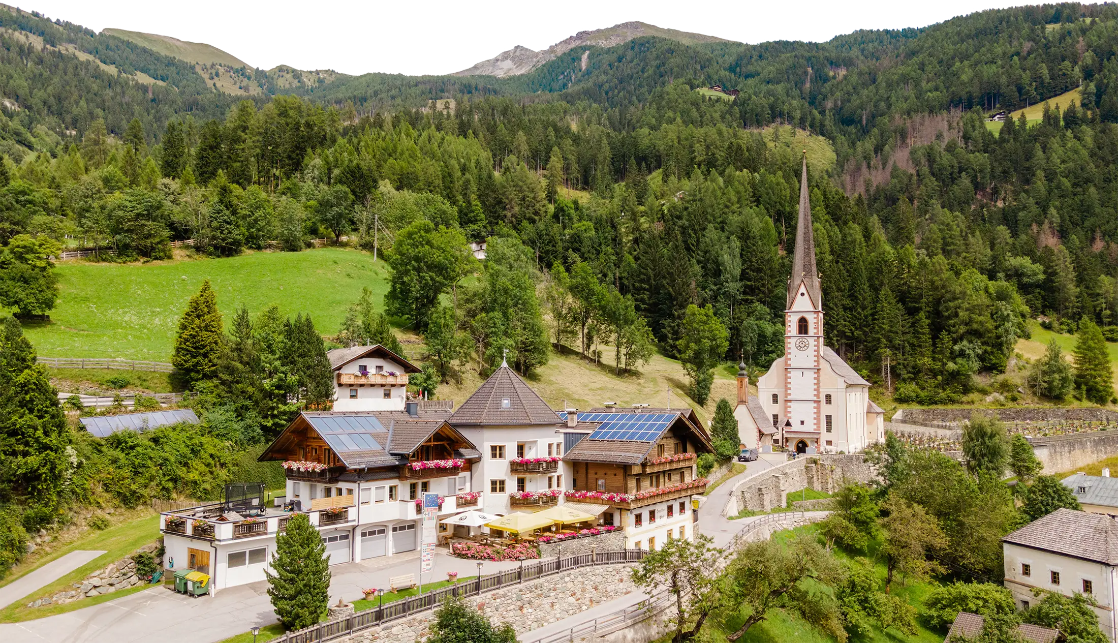 Luftaufnahme des Sagritzer Wirt in Döllach, eingebettet in grüne Berglandschaft, mit angrenzender Kirche und umliegenden Wiesen und Wäldern.